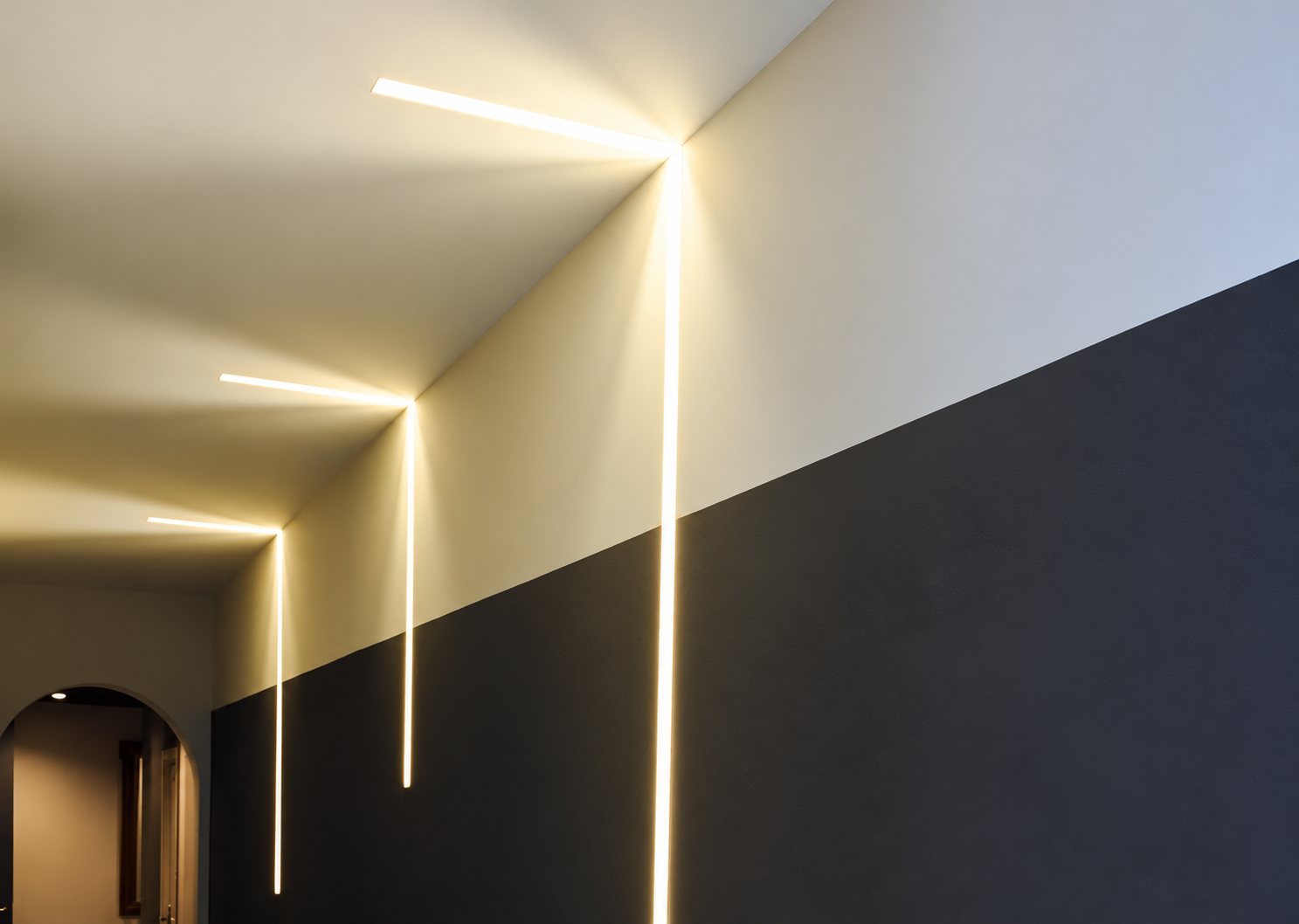 Un couloir aux murs blancs et gris anthracite est éclairé par de fines lignes de lumière encastrées qui descendent du plafond le long du mur, avec une arche au fond à gauche.