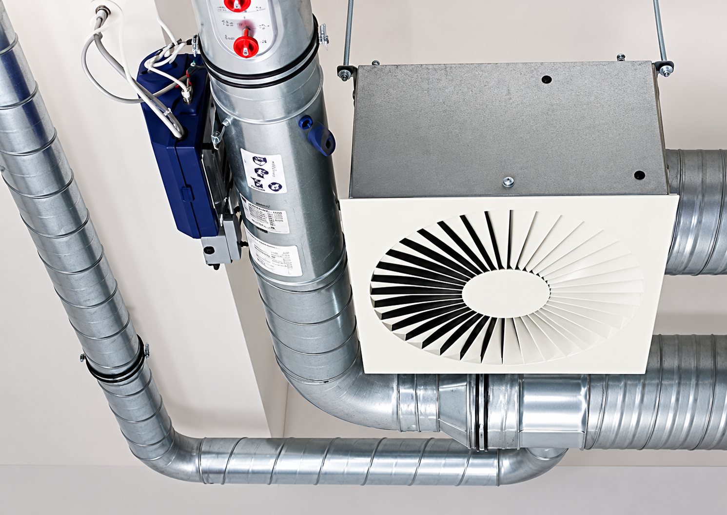 Un diffuseur d'air carré à grille circulaire est installé au plafond, relié à des gaines de ventilation métalliques et à un caisson avec câblage.