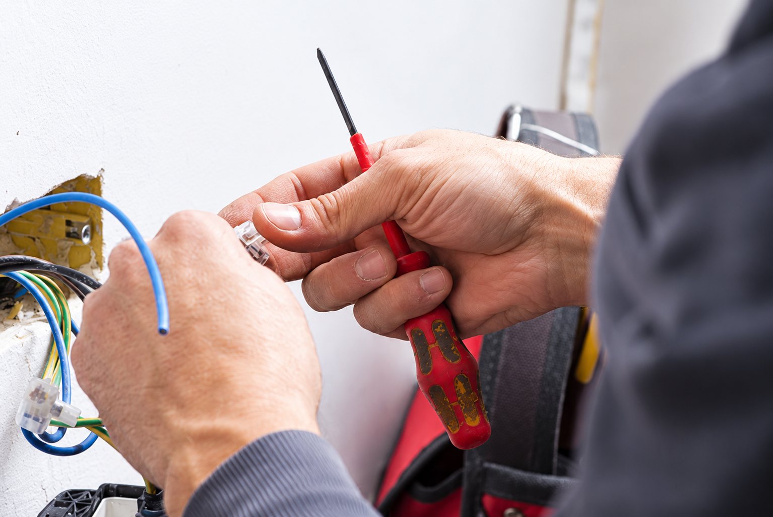 Les mains d'un électricien tiennent un tournevis rouge à côté de câbles électriques bleus et de connecteurs fixés à un mur.