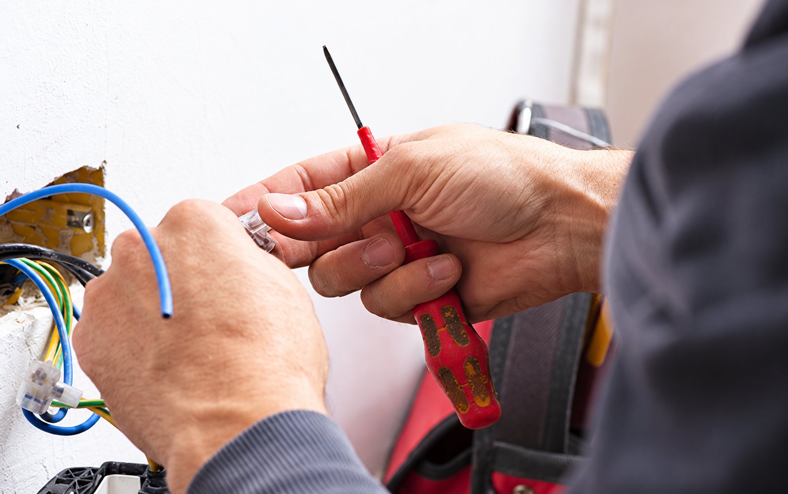 Gros plan sur les mains d'un électricien qui tient un tournevis à manche rouge près de fils électriques bleus et d'une boîte d'encastrement murale.