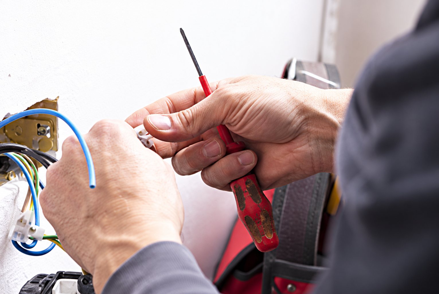 Gros plan sur les mains d'un électricien qui utilise un tournevis à manche rouge près de câbles bleus sur une installation électrique.