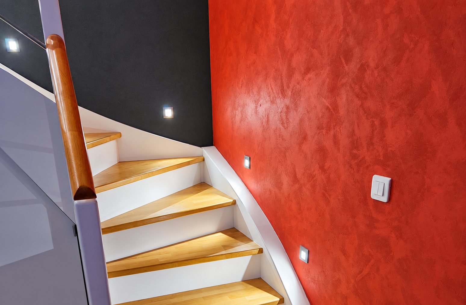 Escalier tournant aux marches en bois clair et contremarches blanches, bordé d'un mur rouge à effet patiné et d'un mur gris foncé, avec petites lumières encastrées.