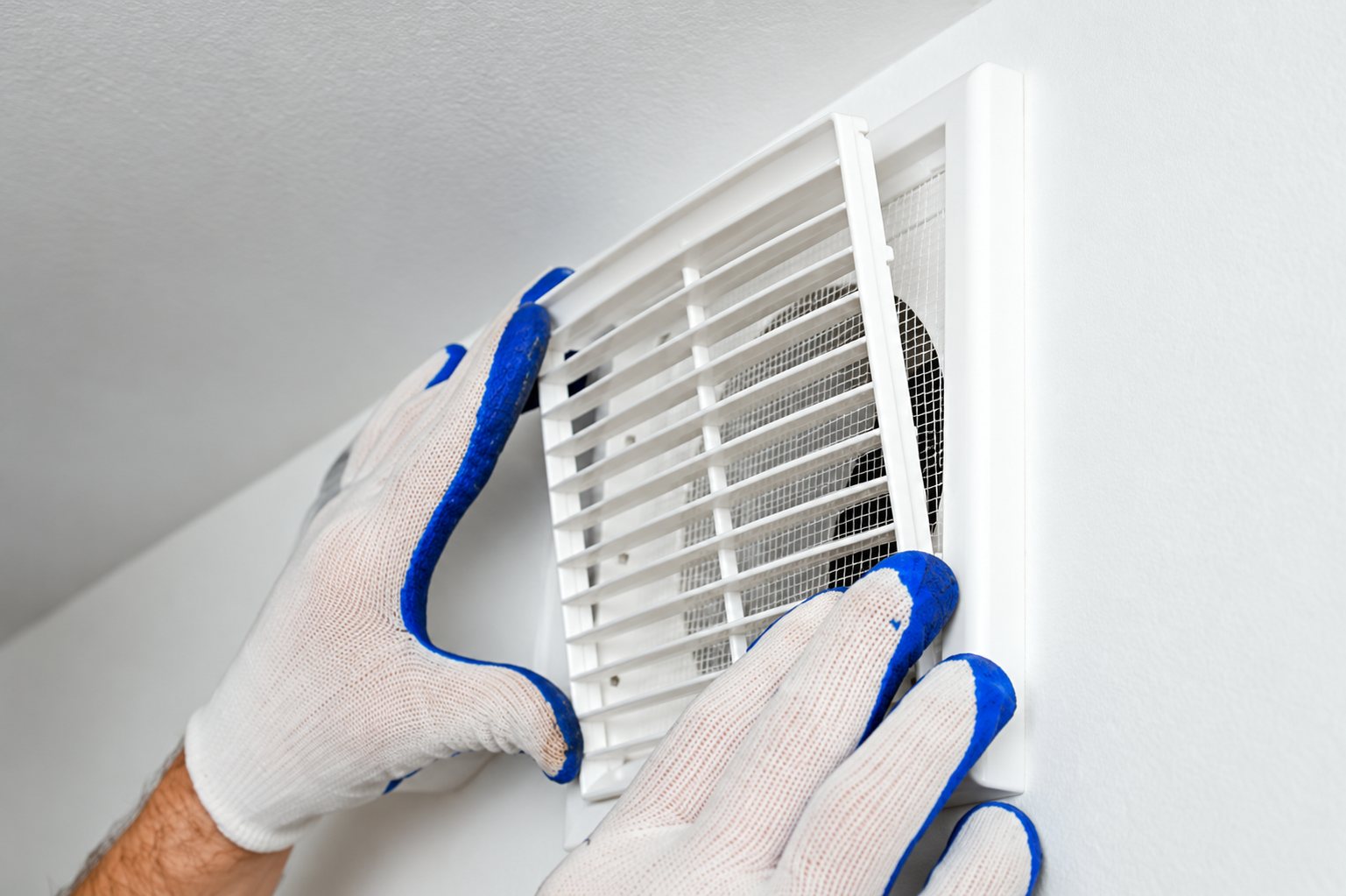 Deux mains portant des gants de travail blancs et bleus fixent une grille de ventilation blanche sur un mur intérieur.