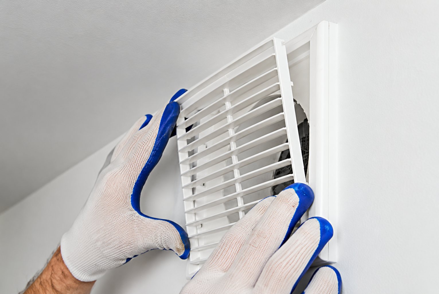 Deux mains portant des gants blancs et bleus posent une grille de ventilation blanche sur un mur clair.