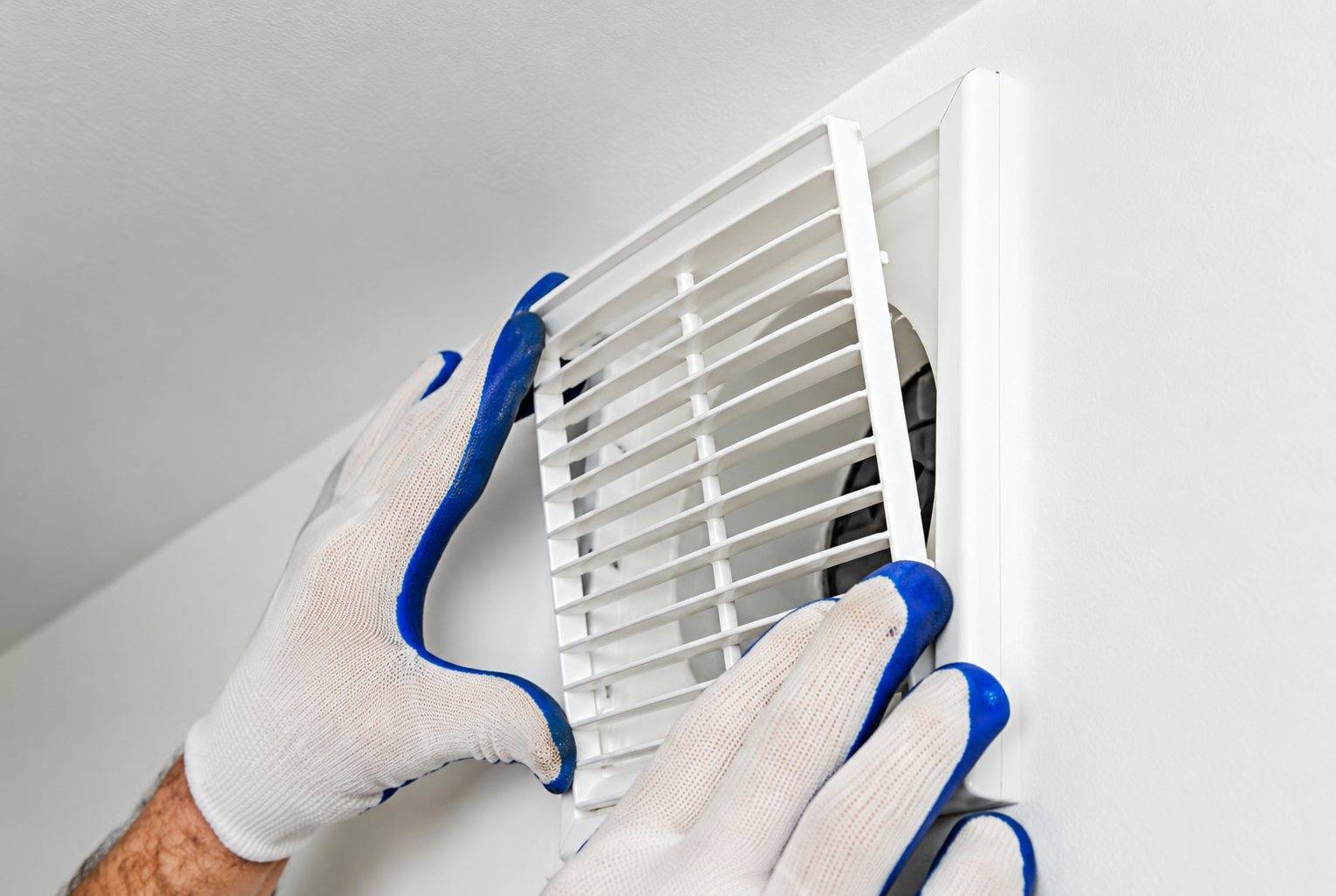 Deux mains portant des gants blancs et bleus posent une grille de ventilation blanche sur un mur clair.