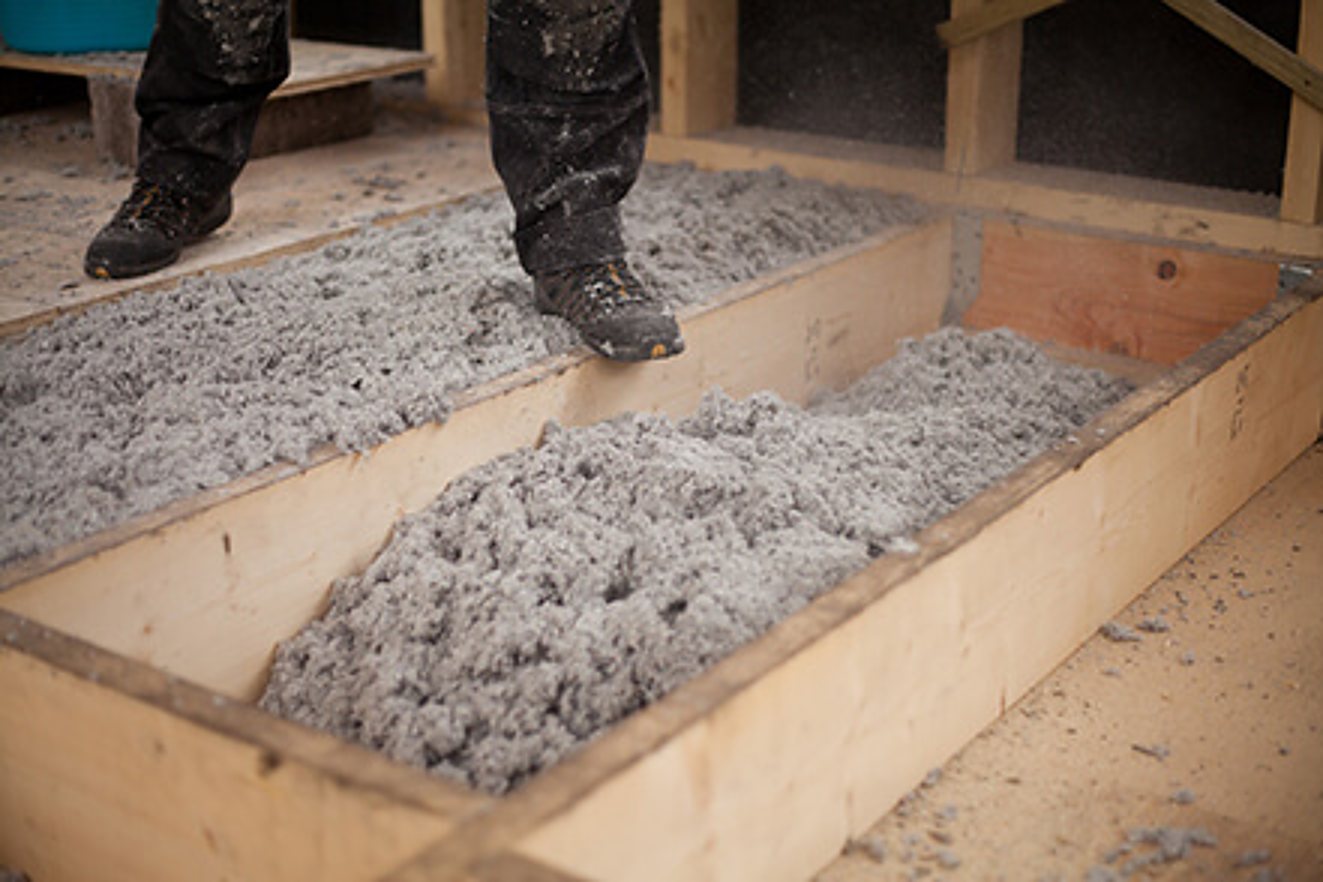Un ouvrier en chaussures de chantier se tient au-dessus de caissons en bois remplis d'isolant en ouate de cellulose grise.