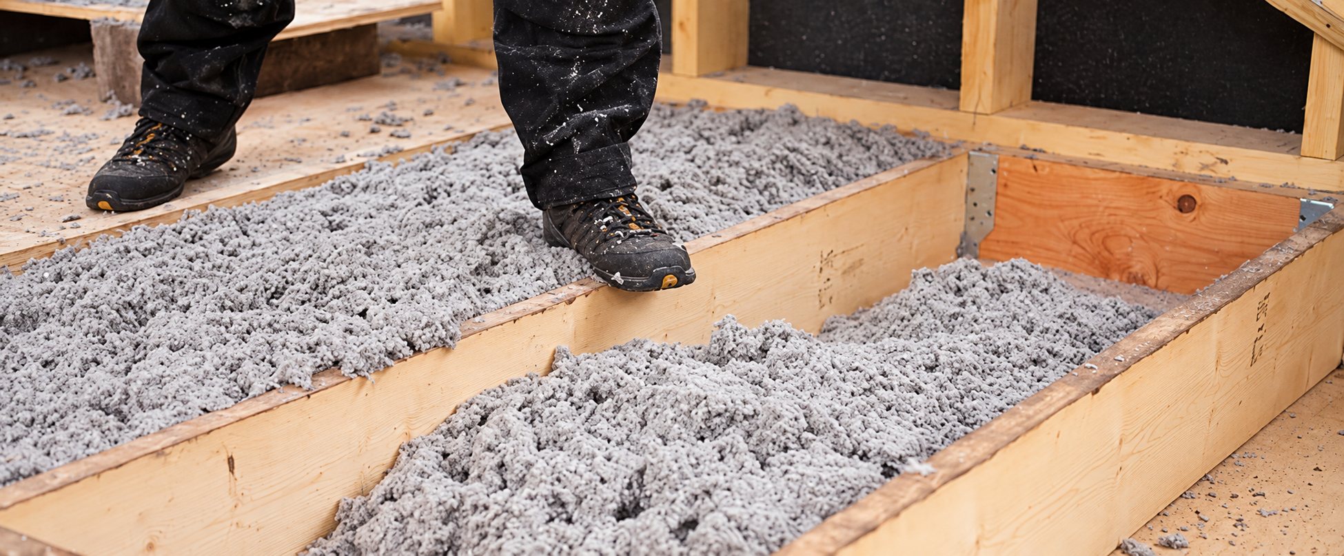 Un ouvrier en chaussures de chantier boueuses se tient au-dessus de solives en bois remplies d'isolant en ouate de cellulose grise.