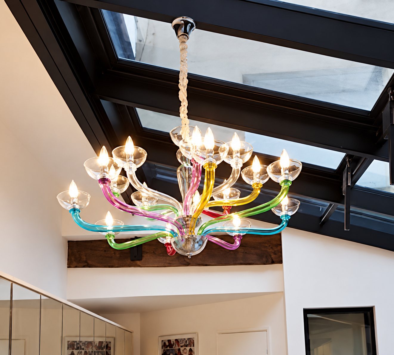 Un lustre moderne aux bras en verre multicolores et aux ampoules allumées est suspendu sous une grande verrière dans une pièce claire.