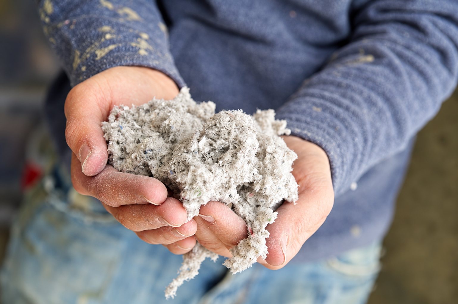 Une personne en pull bleu tient entre ses mains une poignée de ouate de cellulose grise utilisée pour l'isolation.