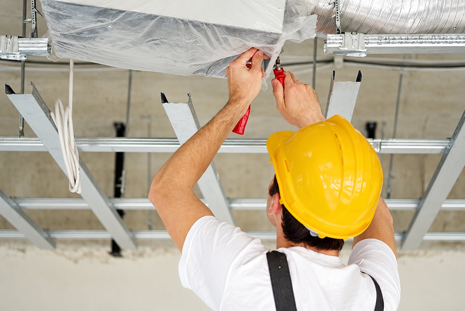 Un ouvrier portant un casque de chantier jaune travaille au plafond, au milieu de rails métalliques et d'une gaine isolée, avec un outil rouge à la main.