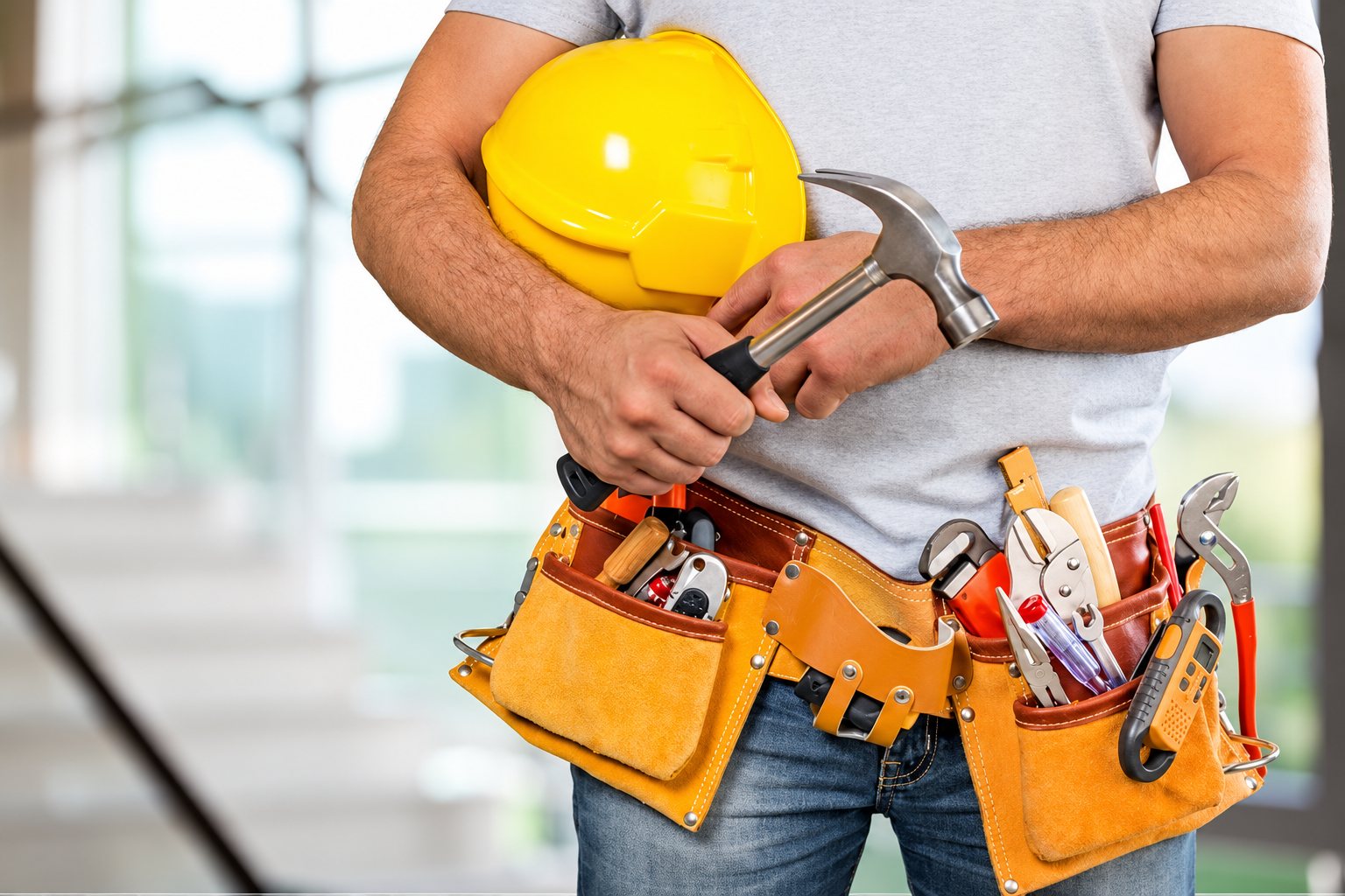 Un ouvrier en t-shirt gris et jean tient un casque de chantier jaune et un marteau, avec une ceinture en cuir remplie d'outils autour de la taille.