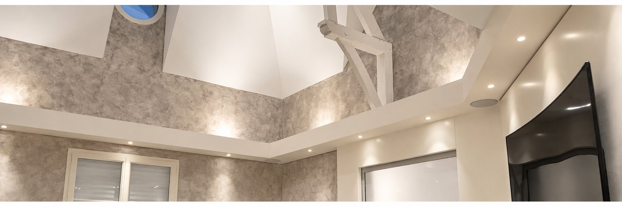 Intérieur aux tons clairs avec un plafond tendu brillant à plusieurs niveaux, des spots encastrés, des poutres apparentes et des murs à l'effet patiné.
