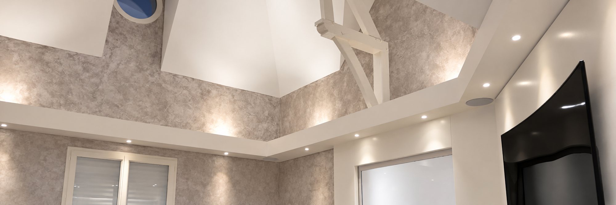 Intérieur clair avec un plafond tendu brillant à plusieurs niveaux, des spots encastrés, des poutres blanches et des murs à l'effet patiné.