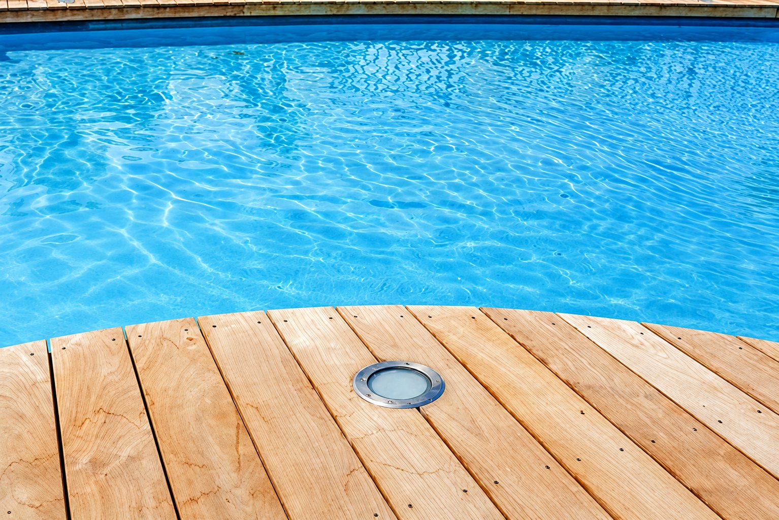 Une terrasse en lames de bois arrondie borde une piscine à l'eau bleue, avec un spot lumineux encastré dans le bois.