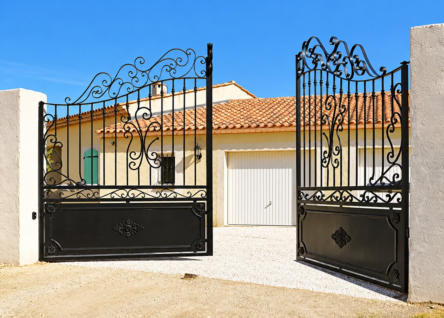 Un portail à deux battants en fer forgé noir aux motifs en volutes, grand ouvert sur une allée de gravier menant à une maison aux murs jaunes et au toit de tuiles.