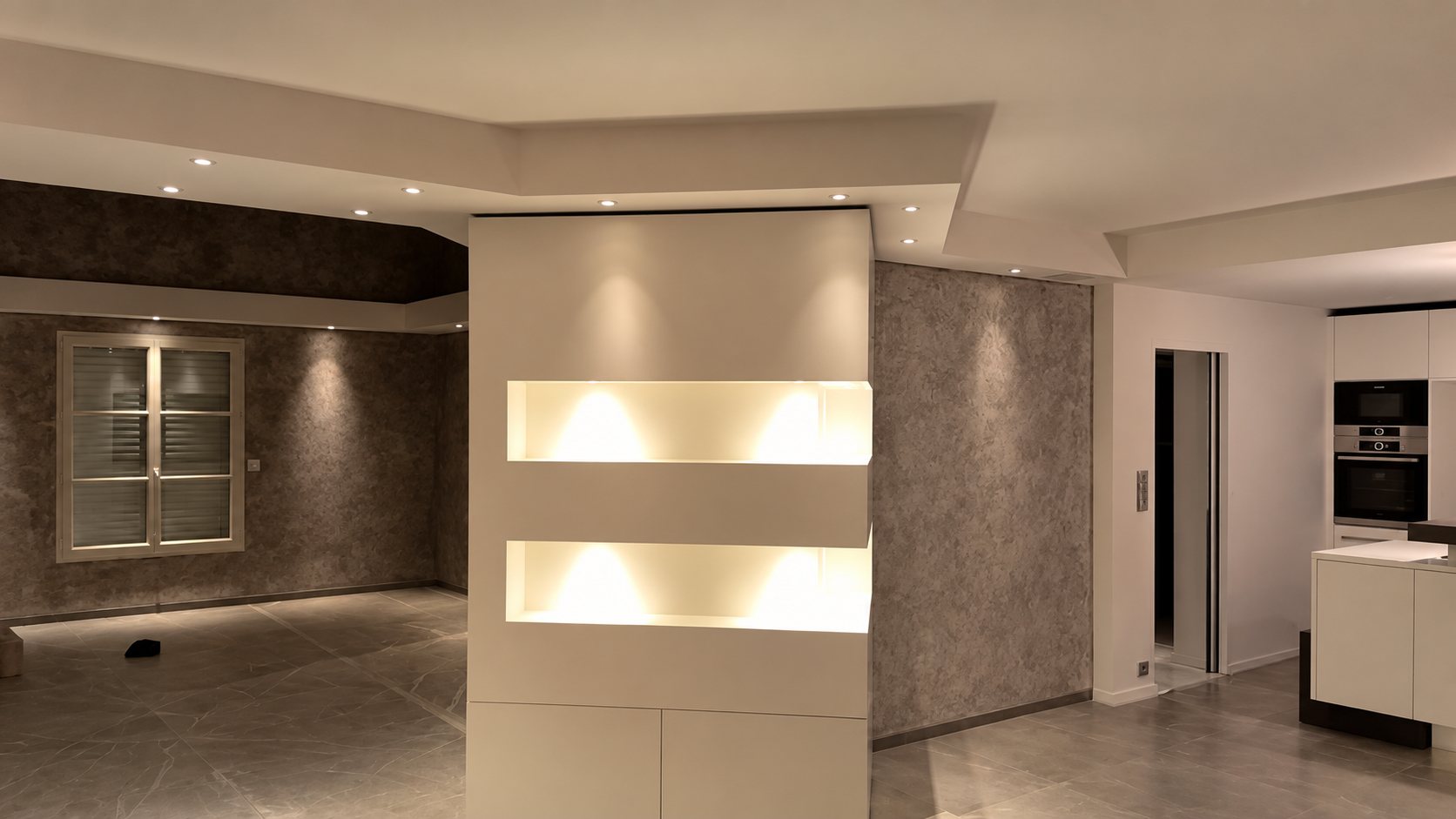 Pièce de vie rénovée avec une cloison blanche à niches éclairées, un faux plafond à spots encastrés, un mur à effet patiné et une cuisine ouverte à droite.