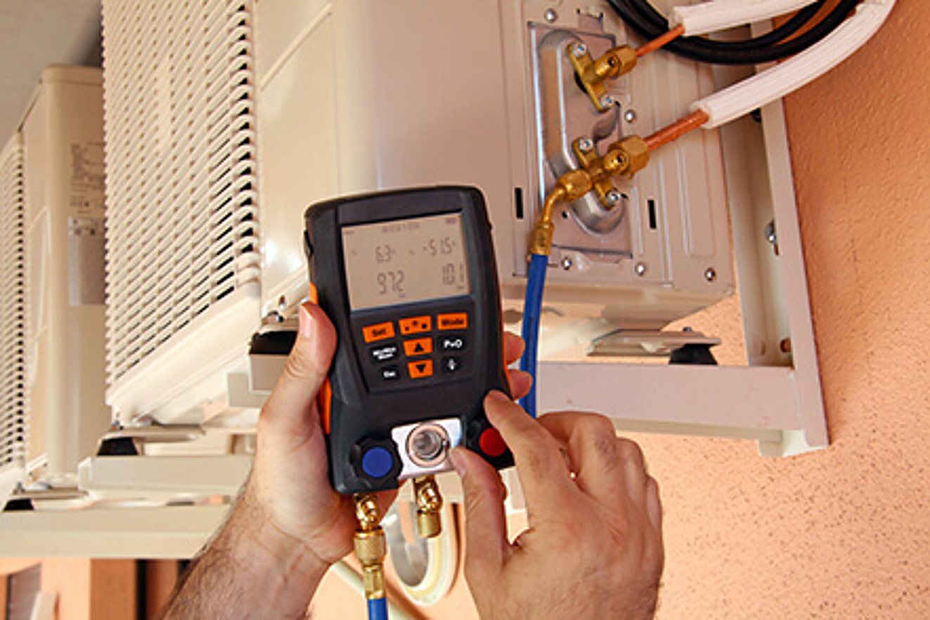 Les mains d'un technicien tiennent un manomètre numérique branché sur les conduites cuivre d'une unité extérieure de climatisation fixée à un mur orangé.
