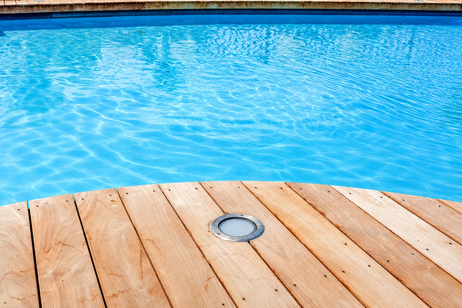 Une terrasse en lames de bois clair épouse la courbe d'une piscine à l'eau bleue, avec un spot rond encastré dans le bois.